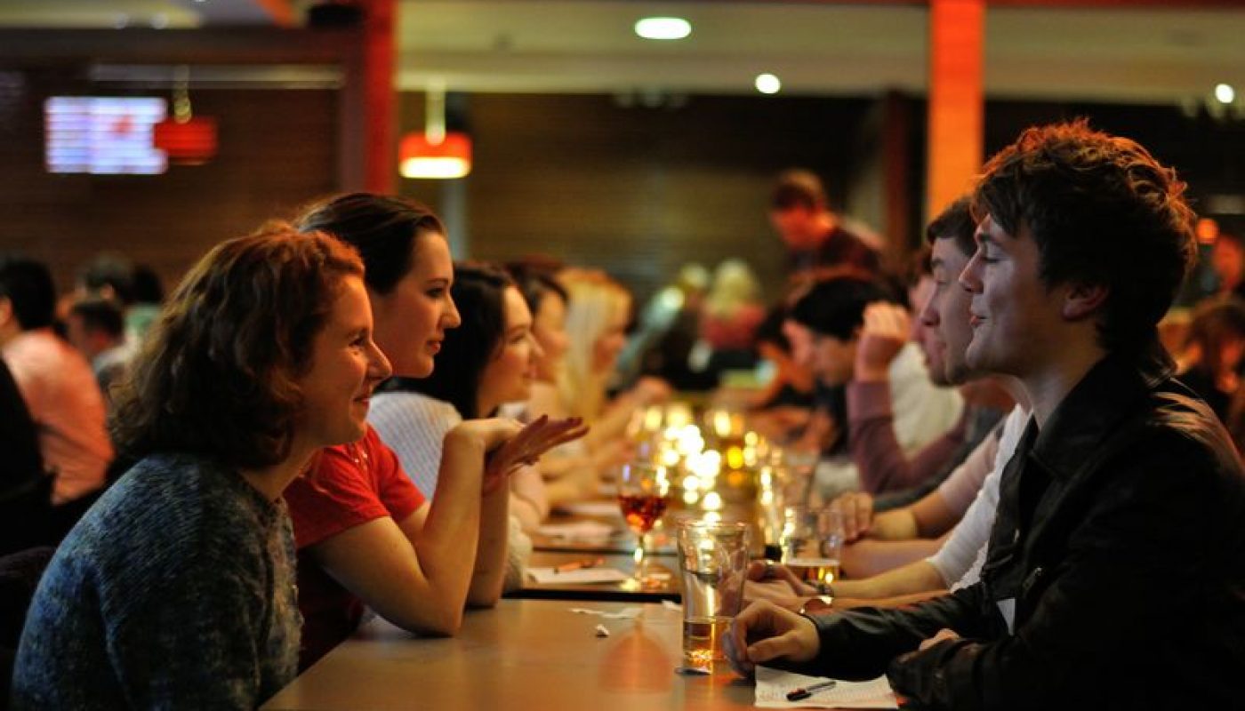 a row of women and row of men sitting in front of each other with wine glasses on top of the table between them