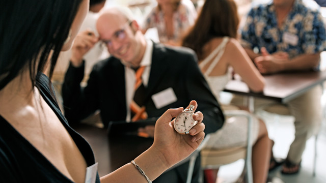 a woman holding a timer and group of people by partners seated facing each other for an speed dating event